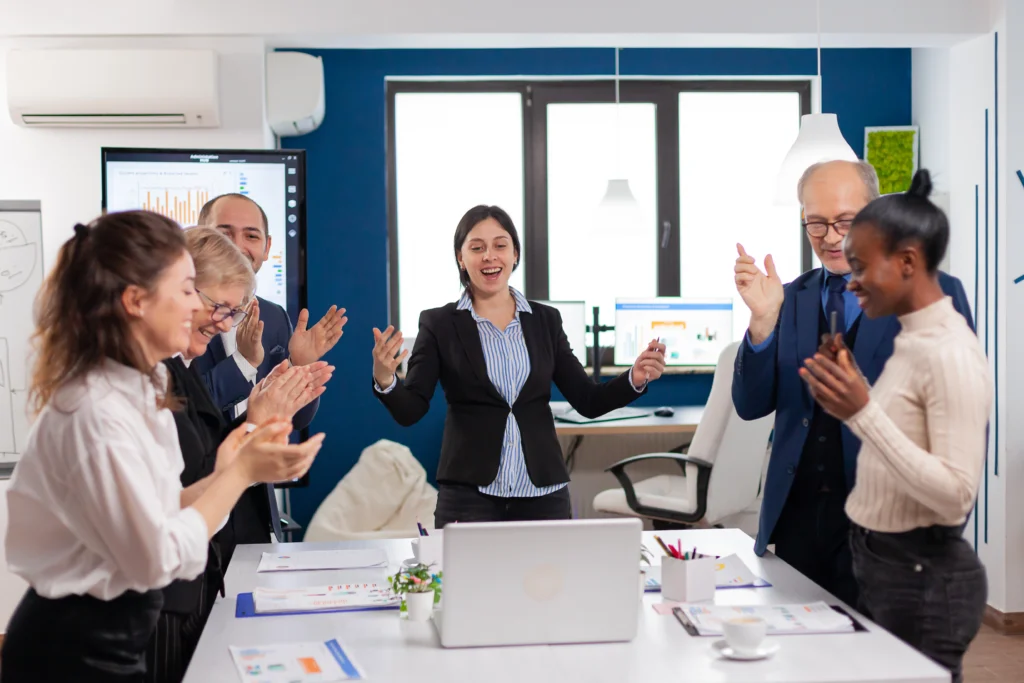 A diverse team of business professionals celebrating and applauding together in a meeting room with performance charts visible on a screen behind them, representing the shared success and measurable growth outcomes that come from experience-led improvement strategies.