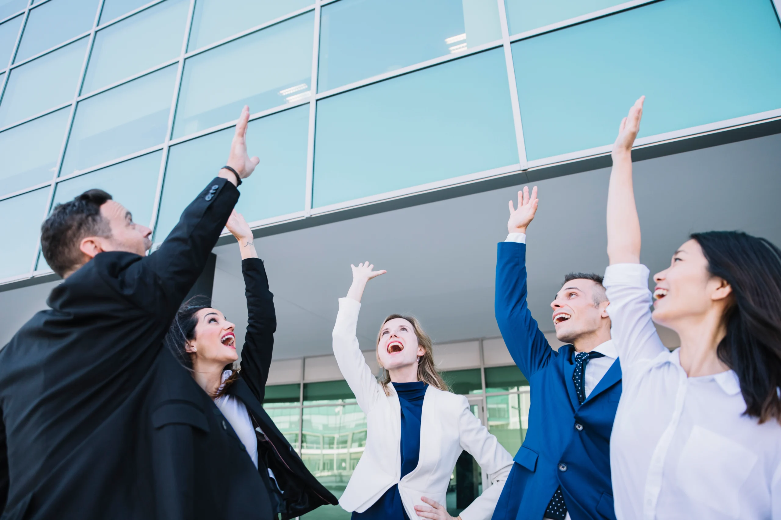 A diverse team of business professionals celebrating together outdoors, raising their hands and smiling in front of a modern office building, symbolising shared success and organisational growth.