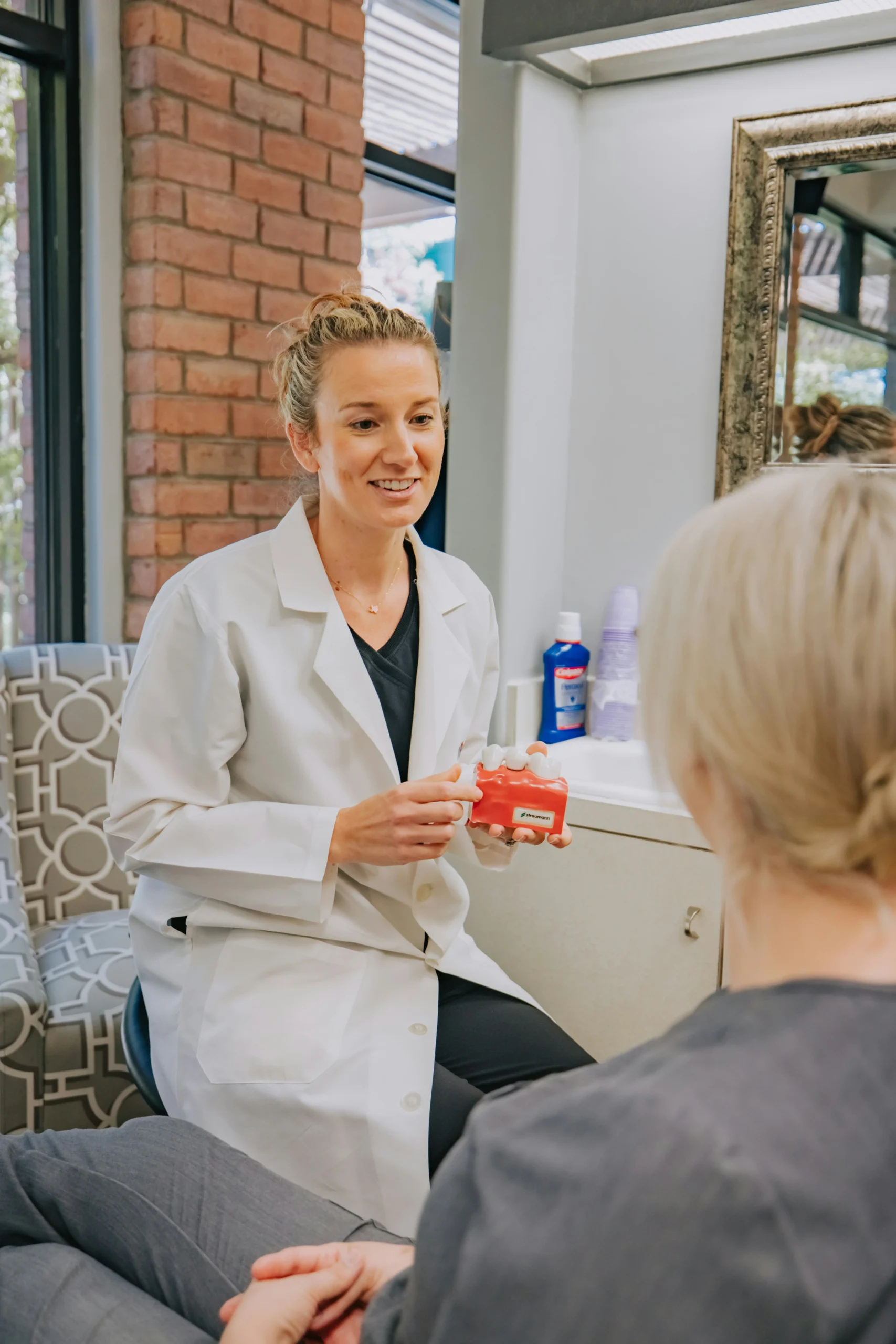 A healthcare clinician smiling and explaining a dental or medical model to a patient during a clinic consultation, representing the attentive, knowledge-driven care that improves patient experience, boosts NPS, and increases clinic bookings.