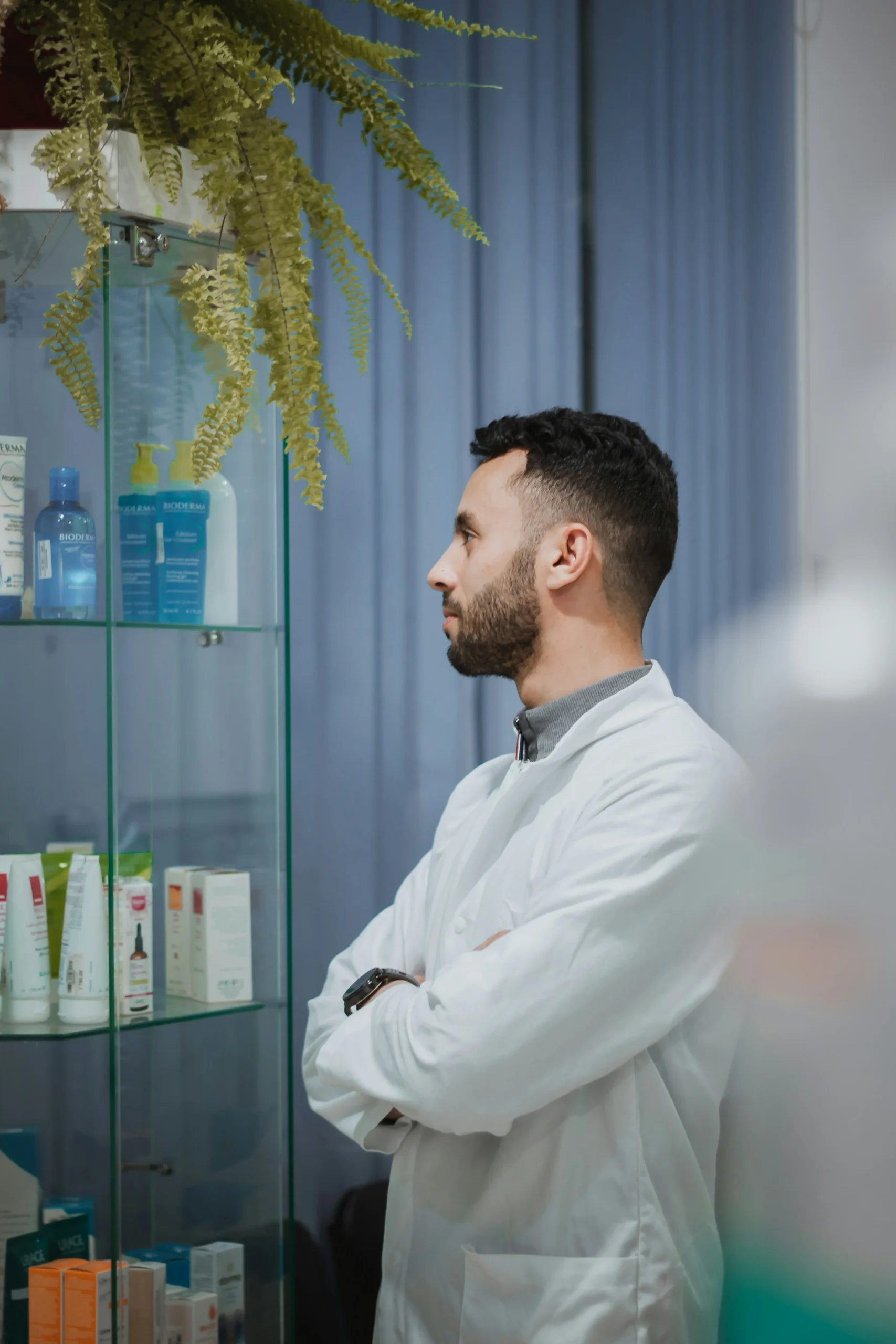 A confident healthcare professional in a white coat standing with arms crossed beside a display of skincare and health products, representing the product knowledge and service standards that drive customer advocacy and pharmacy sales growth.