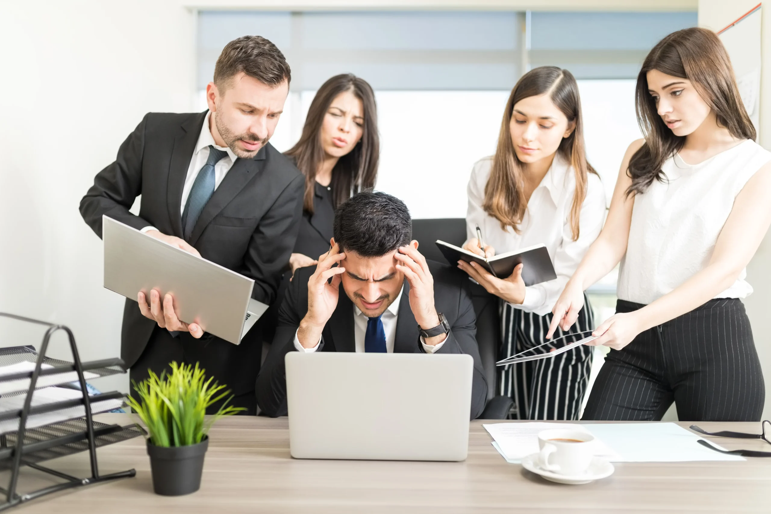 A stressful office scene where a central man in a suit, sitting at a desk with his head in his hands, is overwhelmed by four demanding colleagues.