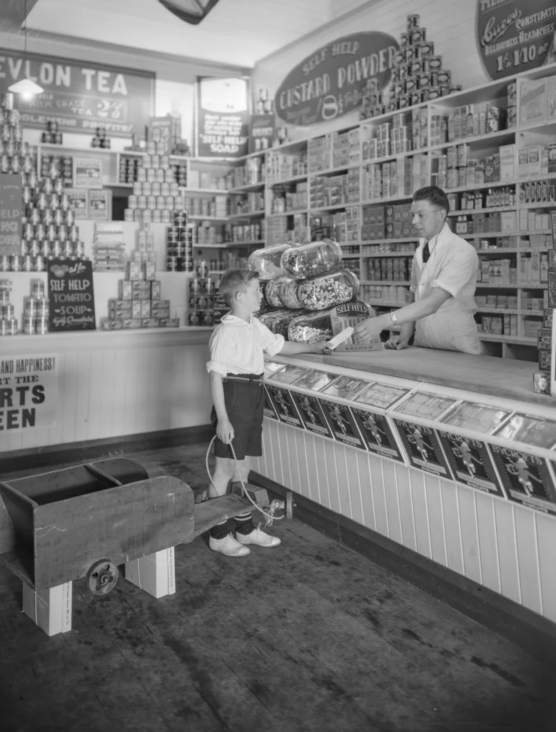 A vintage black and white photograph of a shopkeeper serving a young customer at a traditional grocery store counter, evoking the origins of personalised retail service and the enduring importance of the customer experience.