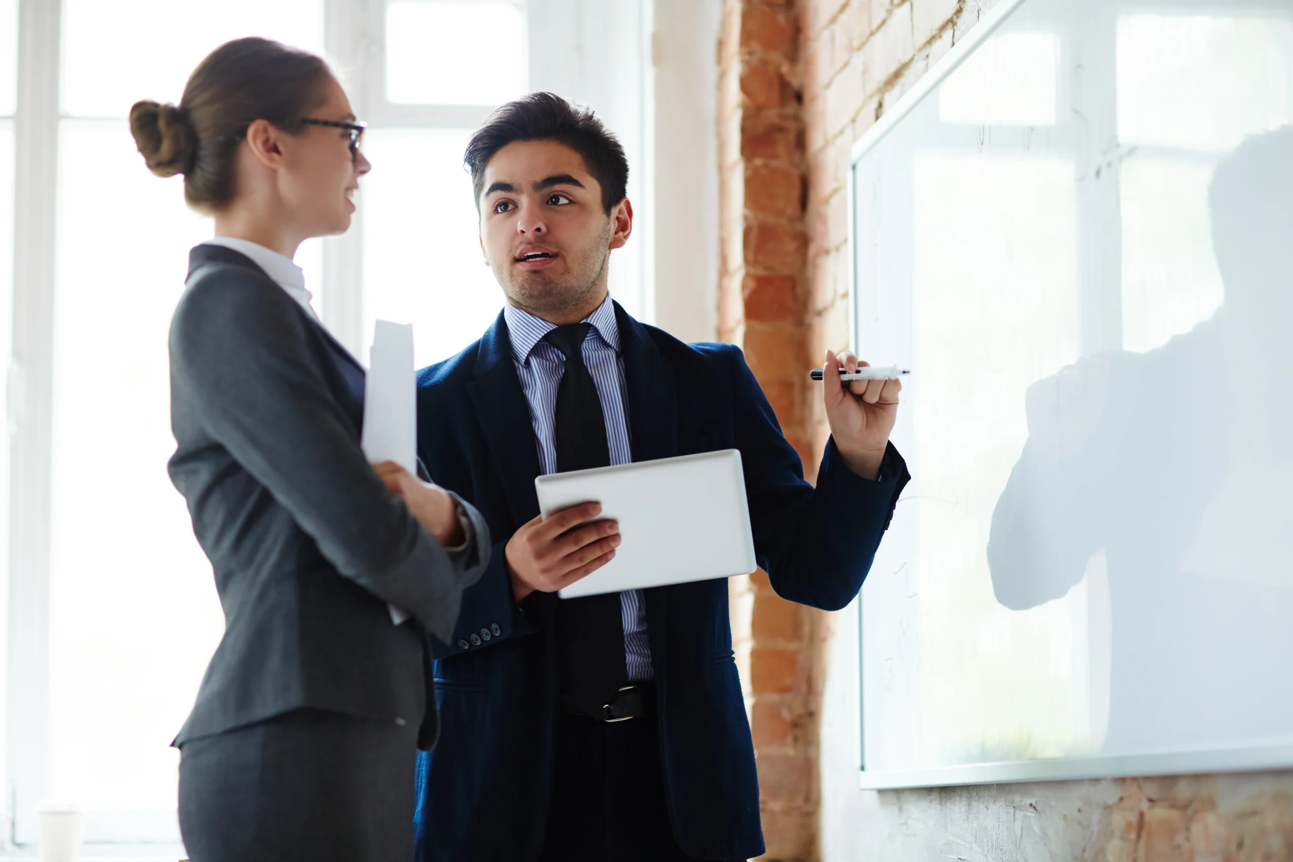 Two business professionals in a focused discussion beside a whiteboard, one holding a tablet and pen, representing the action planning, coaching conversations, and performance improvement strategies that Feedback ASAP enables for frontline teams.
