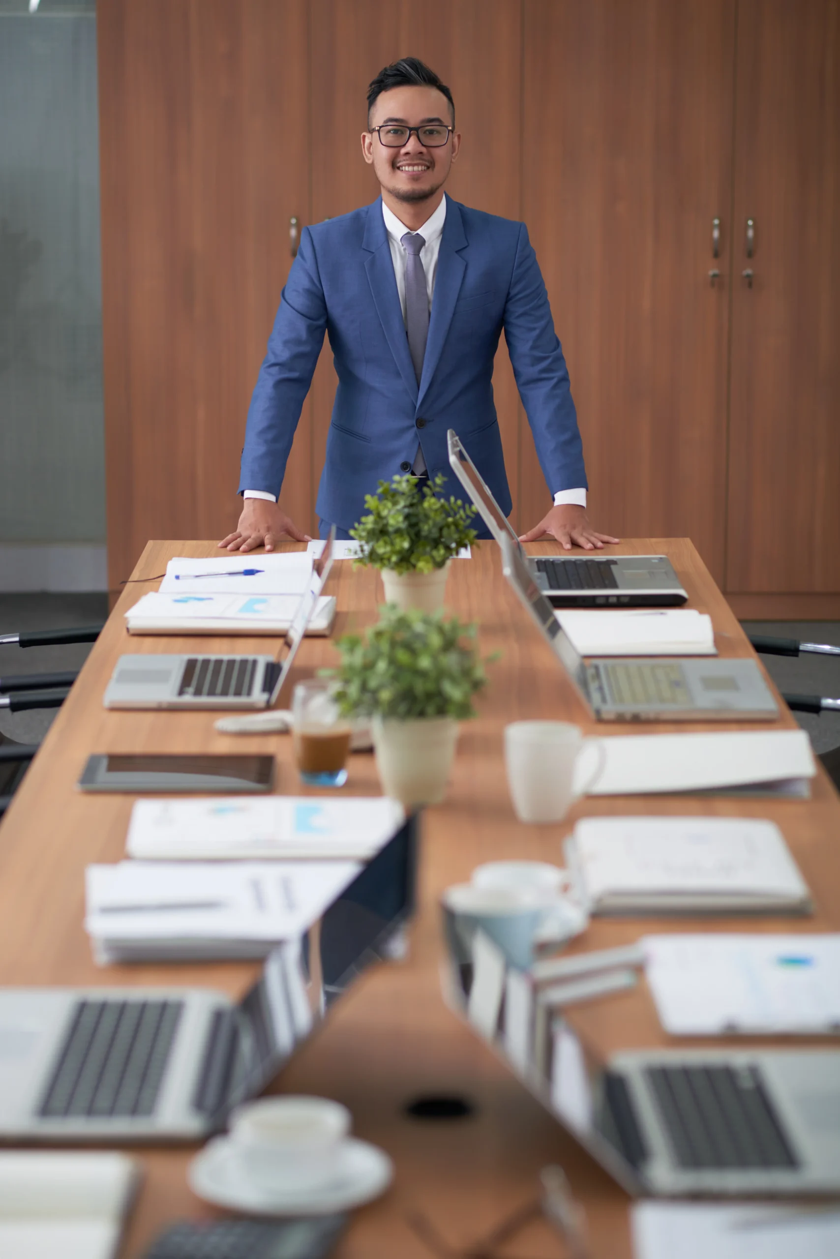 A confident business leader in a blue suit standing at a meeting room table with laptops and documents, representing the executive accountability and results-focused leadership that drives sustainable growth through Feedback ASAP's improvement ecosystem.